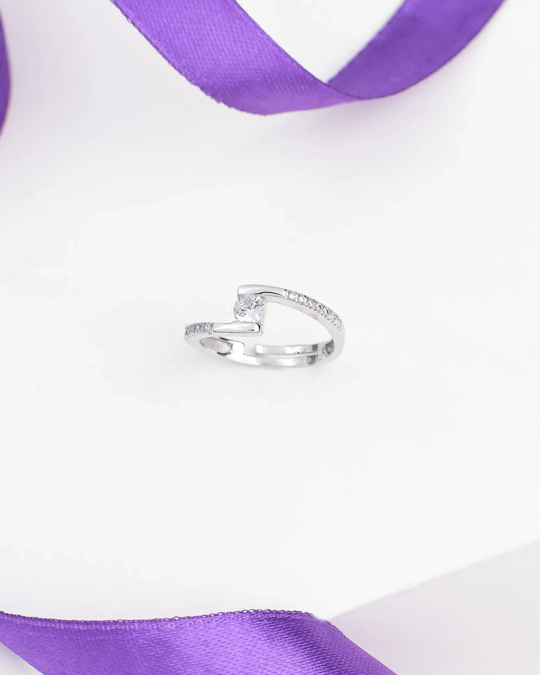The Solitaire CZ Promise Ring in Demifine Silver by Chandrani Pearls features a round central CZ stone and small side stones, shown on a white surface with a purple ribbon in the background.