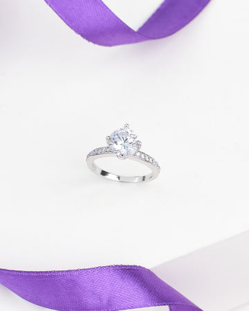 The Classic Demifine Silver Ring with Romantic CZ Center by Chandrani Pearls features a round clear stone and sparkling accents on the band, displayed elegantly on a white surface with a purple ribbon—a timeless jewelry piece.