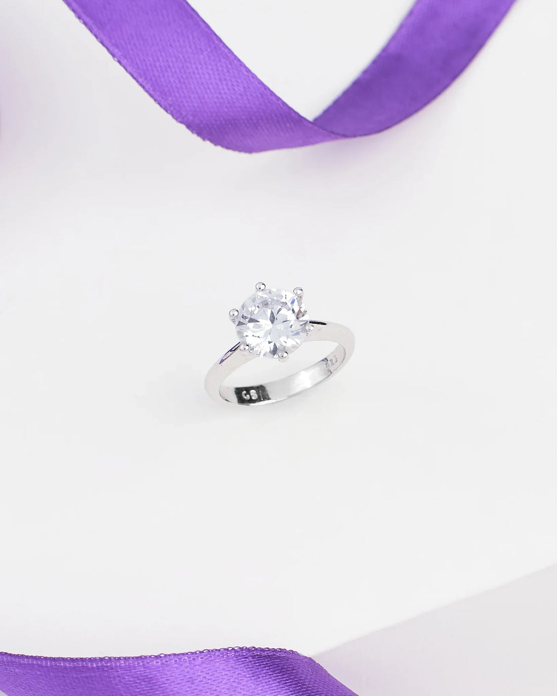 The Elegant Solitaire CZ Ring in Demifine Sterling Silver by Chandrani Pearls is showcased on a white surface with a purple ribbon in the background.
