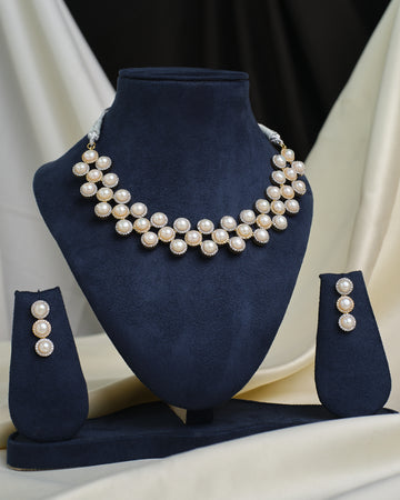 The CZ Infinity Necklace Set by Chandrani Pearls features a pearl necklace, matching pearl earrings, and a CZ-studded infinity pendant, displayed on a navy blue stand with cream and black fabric in the background.