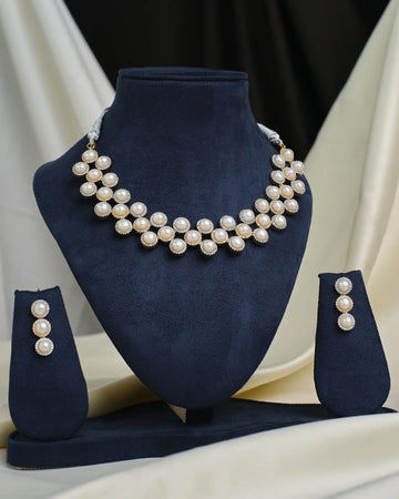 The CZ Infinity Necklace Set by Chandrani Pearls features a pearl necklace, matching pearl earrings, and a CZ-studded infinity pendant, displayed on a navy blue stand with cream and black fabric in the background.