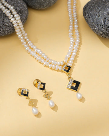 The Chandrani Pearls Regal Golden Grace Pearl & CZ Jewelry Set features a double-strand necklace and matching earrings with gold-tone accents, black geometric details, sparkling CZ stones, and pearl drops. Displayed on beige with gray stones.