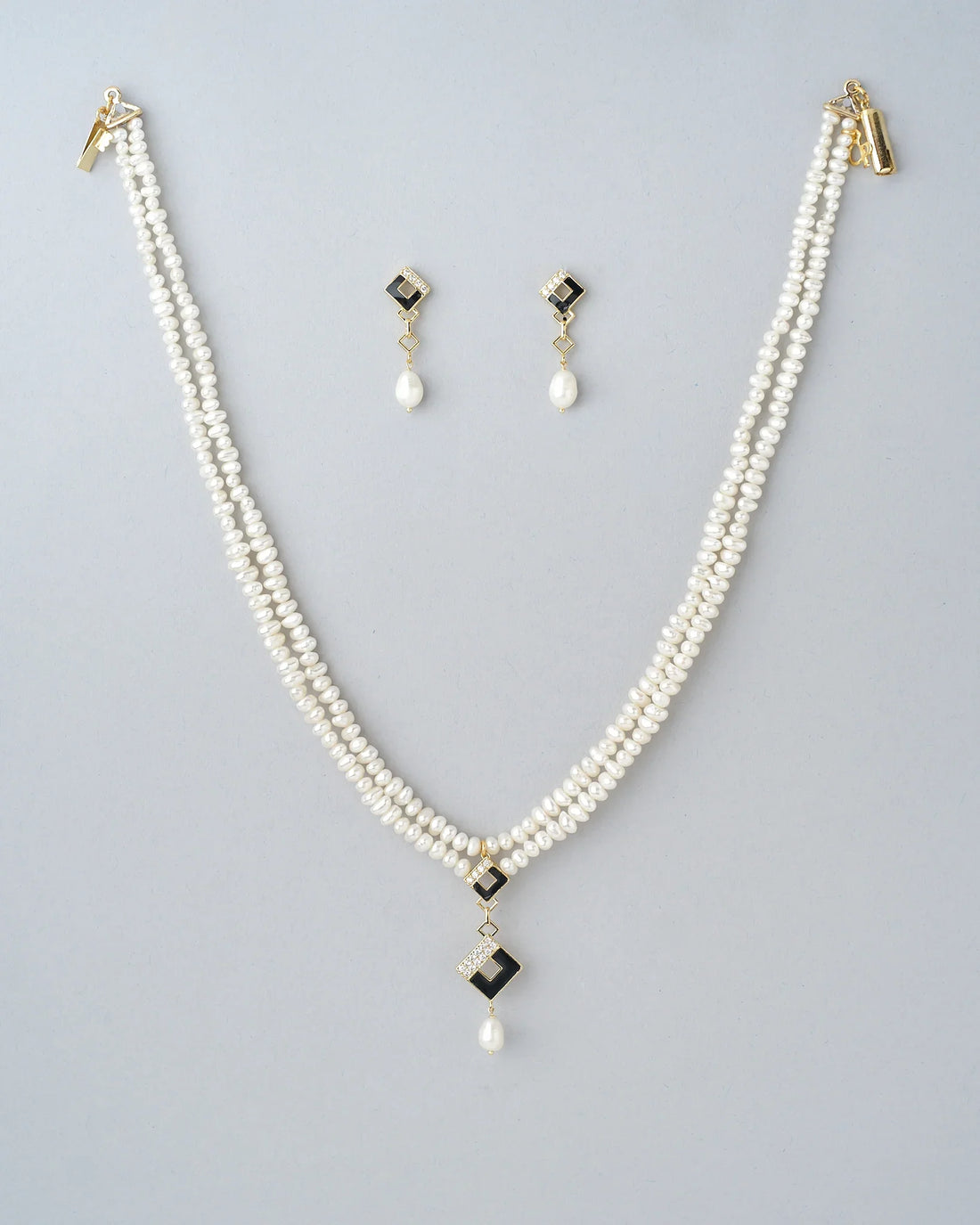 The Chandrani Pearls Regal Golden Grace Pearl & CZ Jewelry Set features a double-strand necklace and matching earrings with gold-tone accents, black geometric details, sparkling CZ stones, and pearl drops. Displayed on beige with gray stones.