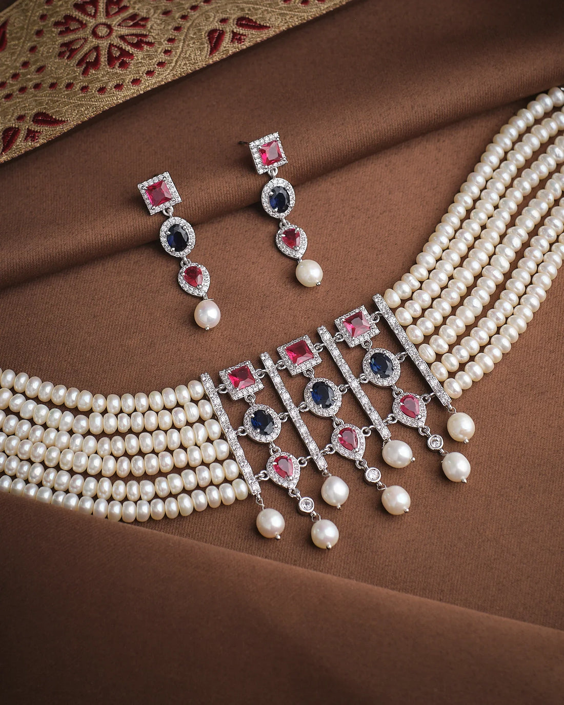 The Empress Pearl & CZ Necklace Set by Chandrani Pearls features layered pearl strands, red, blue, and white stones, CZ accents, and matching earrings—displayed on brown fabric with a decorative border for regal elegance.