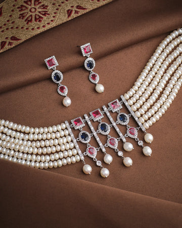 The Empress Pearl & CZ Necklace Set by Chandrani Pearls features layered pearl strands, red, blue, and white stones, CZ accents, and matching earrings—displayed on brown fabric with a decorative border for regal elegance.