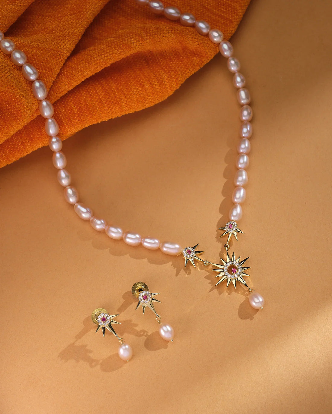 The Zara Gold Layered Set by Chandrani Pearls features a pearl necklace with gold starburst pendants and matching minimalist pearl drop studs, elegantly displayed on a tan surface with orange fabric.