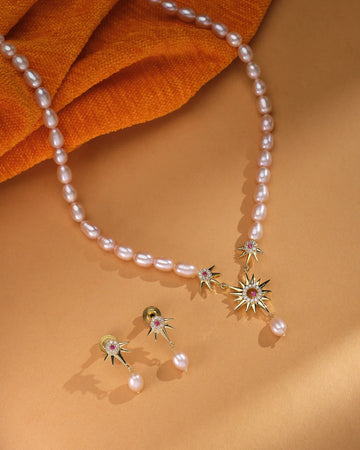 The Zara Gold Layered Set by Chandrani Pearls features a pearl necklace with gold starburst pendants and matching minimalist pearl drop studs, elegantly displayed on a tan surface with orange fabric.