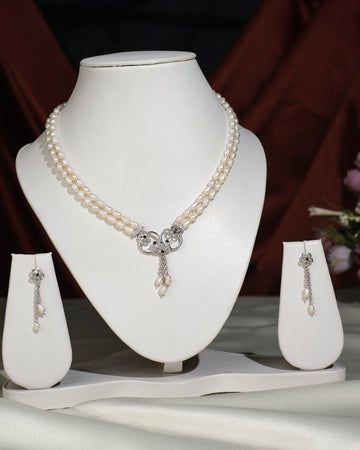 The Trendy Pearl Necklace Set by Chandrani Pearls India features a silver pendant and matching pearl drop earrings, elegantly displayed on white stands with a blurred background.