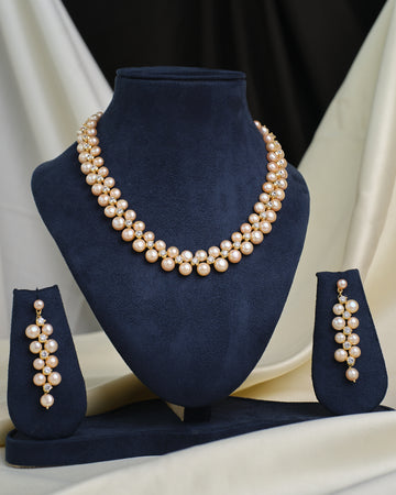 The Royal Mikkimotor Pink CZ Crown Set by Chandrani Pearls, featuring a pearl necklace with a pink CZ crown pendant and matching earrings, is displayed on navy blue velvet stands against a cream and black fabric background.