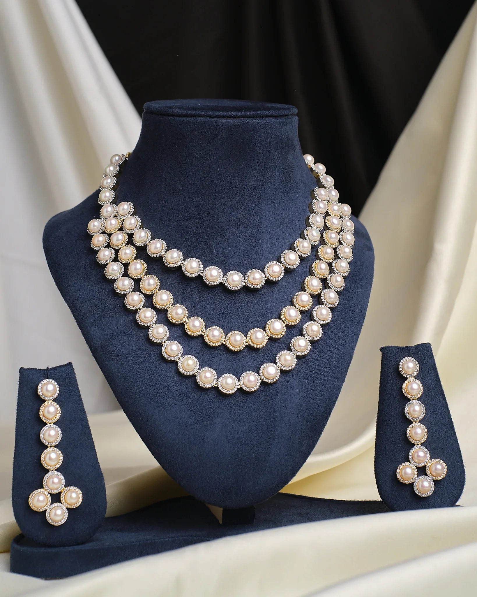 The Glamorous Glow CZ Necklace Set by Chandrani Pearls features a three-layered pearl and CZ necklace with a radiant center stone and matching pearl earrings, displayed on a navy blue stand against a cream and black fabric background.
