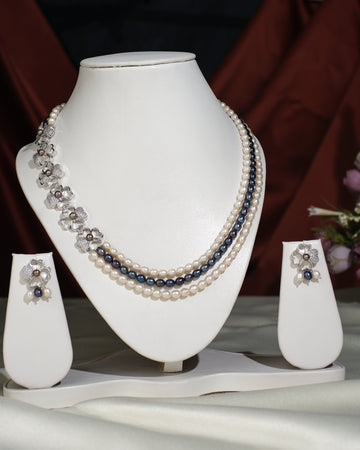 The Florious Side Pearl Necklace Set by Chandrani Pearls India features pearl and gemstone necklaces with floral earrings, elegantly displayed on a white stand against a light cloth background.