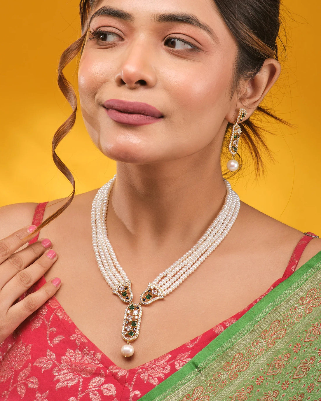 The Queen’s Aura Pearl CZ Necklace Set by Chandrani Pearls is a three-layer pearl necklace with matching earrings, adorned with gold, green, and white CZ stones for a royal look, shown on brown fabric with decorative borders.