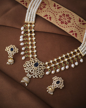 The Royal Celestial Pearl Light Necklace Set by Chandrani Pearls, featuring pearls, blue gemstones, CZ accents, and matching earrings, is displayed on brown fabric with a patterned textile in the background.