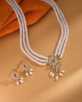 The Golden Bloom Flory Designer Set by Chandrani Pearls features three pearl strands and a floral jeweled pendant with matching earrings, elegantly displayed on a tan surface with orange textured fabric.