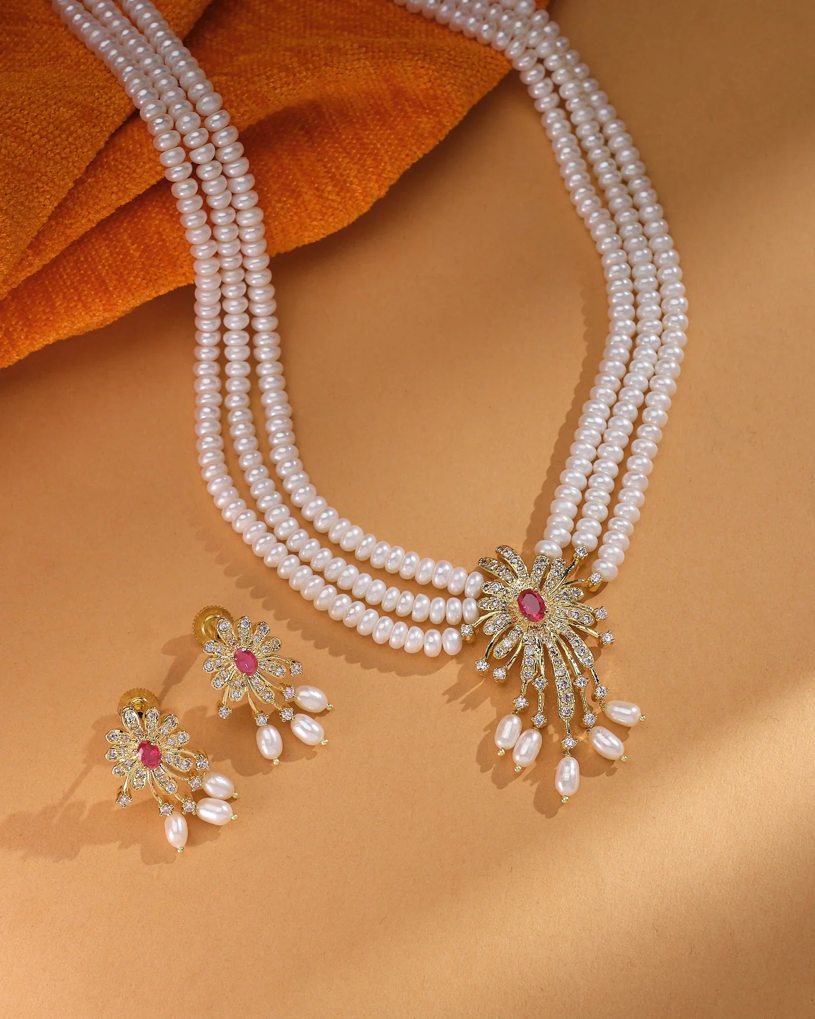 The Golden Bloom Flory Designer Set by Chandrani Pearls features three pearl strands and a floral jeweled pendant with matching earrings, elegantly displayed on a tan surface with orange textured fabric.