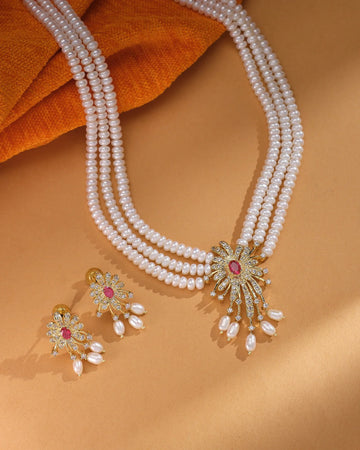 The Golden Bloom Flory Designer Set by Chandrani Pearls features three pearl strands and a floral jeweled pendant with matching earrings, elegantly displayed on a tan surface with orange textured fabric.
