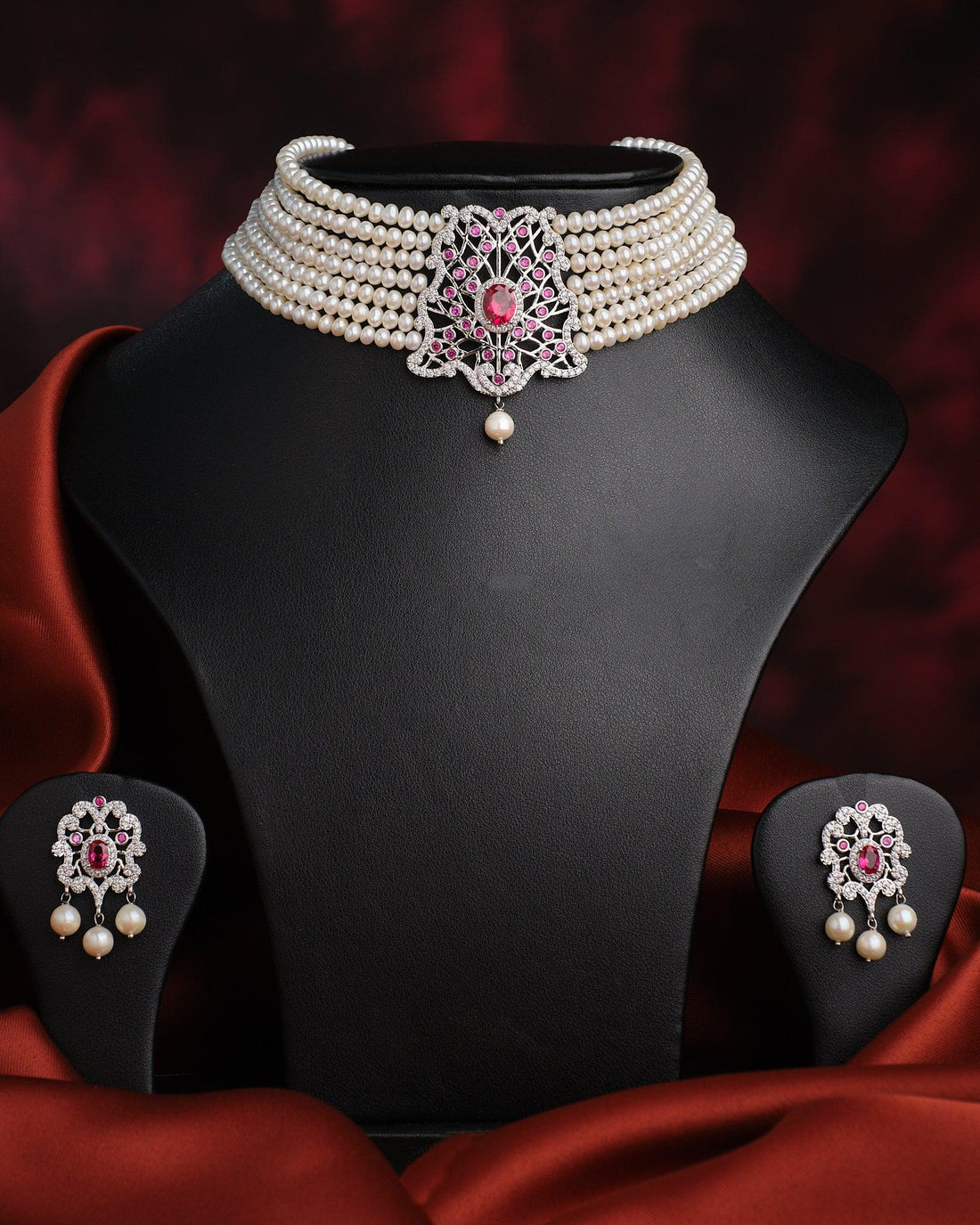 The Somalekha-Modern Yet Traditional Pearl Necklace Set by Chandrani Pearls features a central silver and pink floral pendant with matching earrings, elegantly displayed on a black stand against red satin fabric.