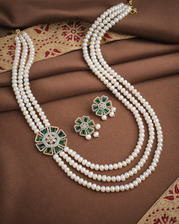 The Noor Floral Side Supreme Pearl CZ Necklace Set by Chandrani Pearls features three layers of premium pearls, green and gold floral pendants, and matching earrings, displayed on brown and gold embroidered fabric.