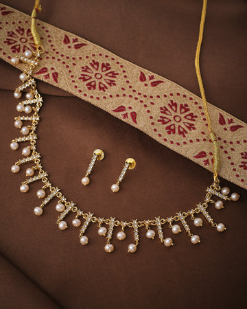 The Pearl & CZ Imperial Layers Necklace Set by Chandrani Pearls, featuring pearl drops, CZ motifs, and matching gold and pearl earrings, is displayed on brown fabric with a gold and red embroidered cloth.