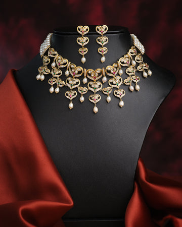 The Dazzling Pearl Necklace Set by Chandrani Pearls features a gold and gemstone necklace with matching earrings, accented with pearls and red fabric on a black bust—ideal for formal occasions.