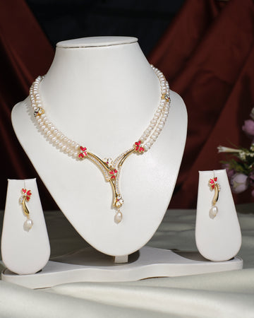 The Floral Enamel Pearl Necklace Set by Chandrani Pearls India features crafted pearls with gold and red accents on a white stand, paired with matching earrings, displayed against a dark draped background.
