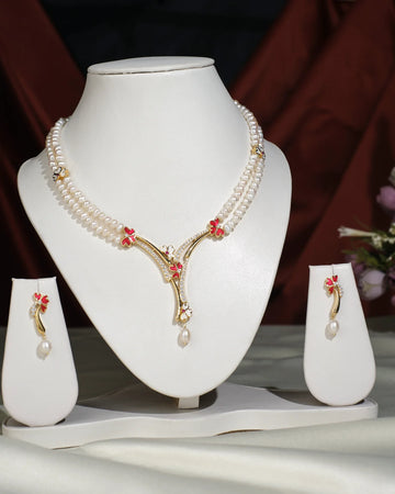 The Floral Enamel Pearl Necklace Set by Chandrani Pearls India features crafted pearls with gold and red accents on a white stand, paired with matching earrings, displayed against a dark draped background.