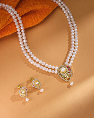 The Celina CZ Pearl Set by Chandrani Pearls—featuring a refined double-strand pearl necklace with a gold and gemstone pendant plus matching pearl and gold earrings—radiates timeless brilliance on a tan background.