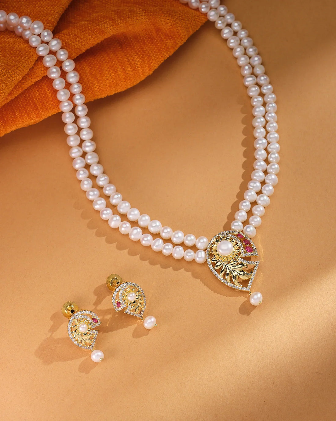 The Celina CZ Pearl Set by Chandrani Pearls—featuring a refined double-strand pearl necklace with a gold and gemstone pendant plus matching pearl and gold earrings—radiates timeless brilliance on a tan background.
