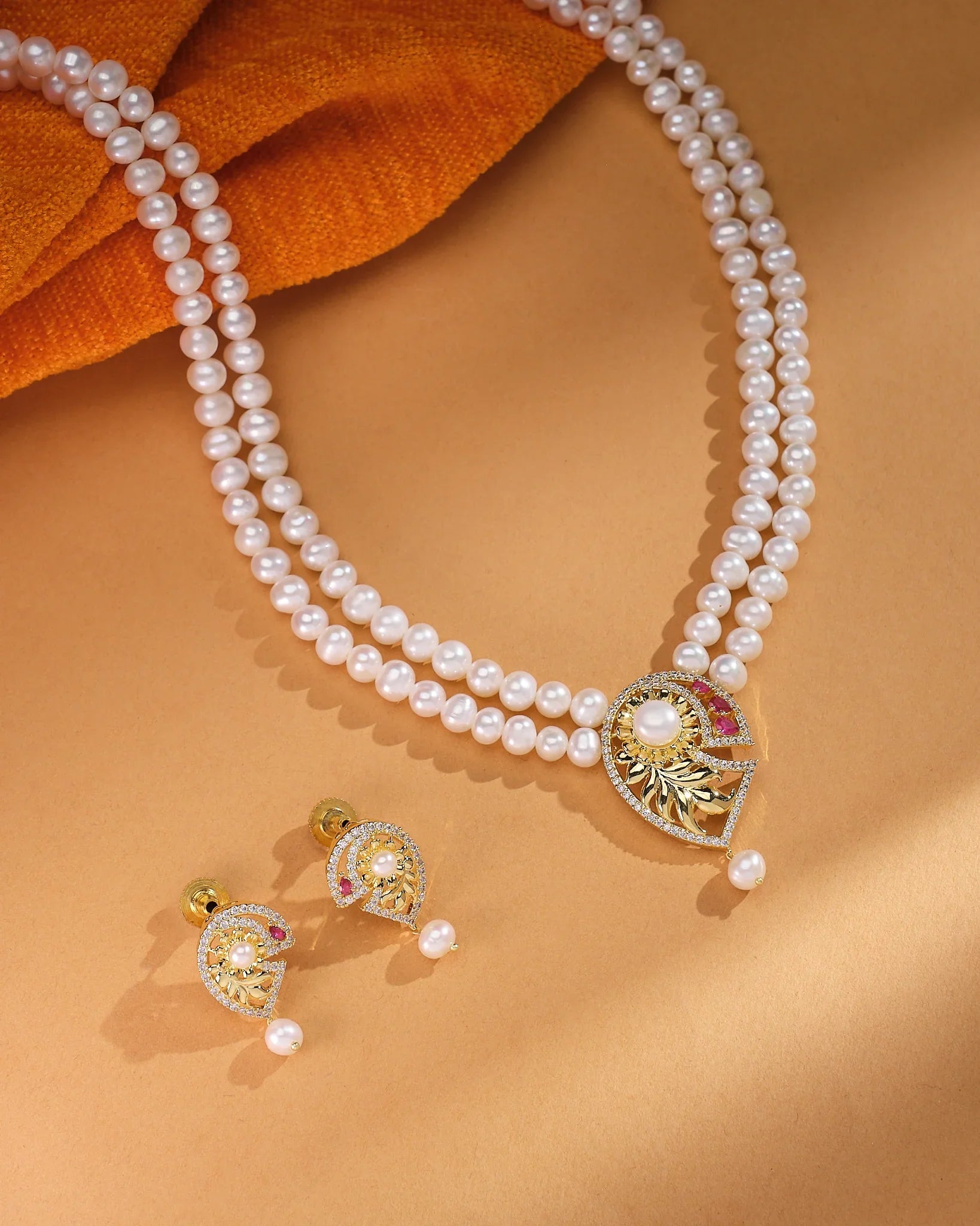 The Celina CZ Pearl Set by Chandrani Pearls—featuring a refined double-strand pearl necklace with a gold and gemstone pendant plus matching pearl and gold earrings—radiates timeless brilliance on a tan background.