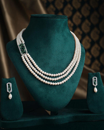 The Sensational Overlapping Shine Pearl Necklace Set by Chandrani Pearls, featuring a three-strand design with green and silver accents, is showcased on a dark green velvet stand, with matching earrings included.