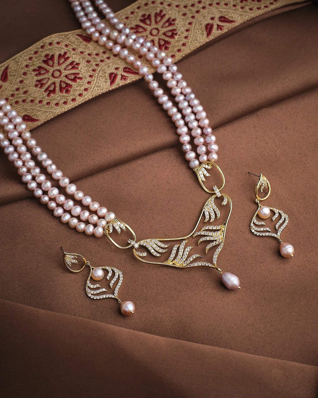 The Regal Pearl Unity Necklace Set by Chandrani Pearls features a three-strand pink pearl necklace with gold, diamond, and CZ accents, matching earrings, and gold-embroidered fabric on a brown background.