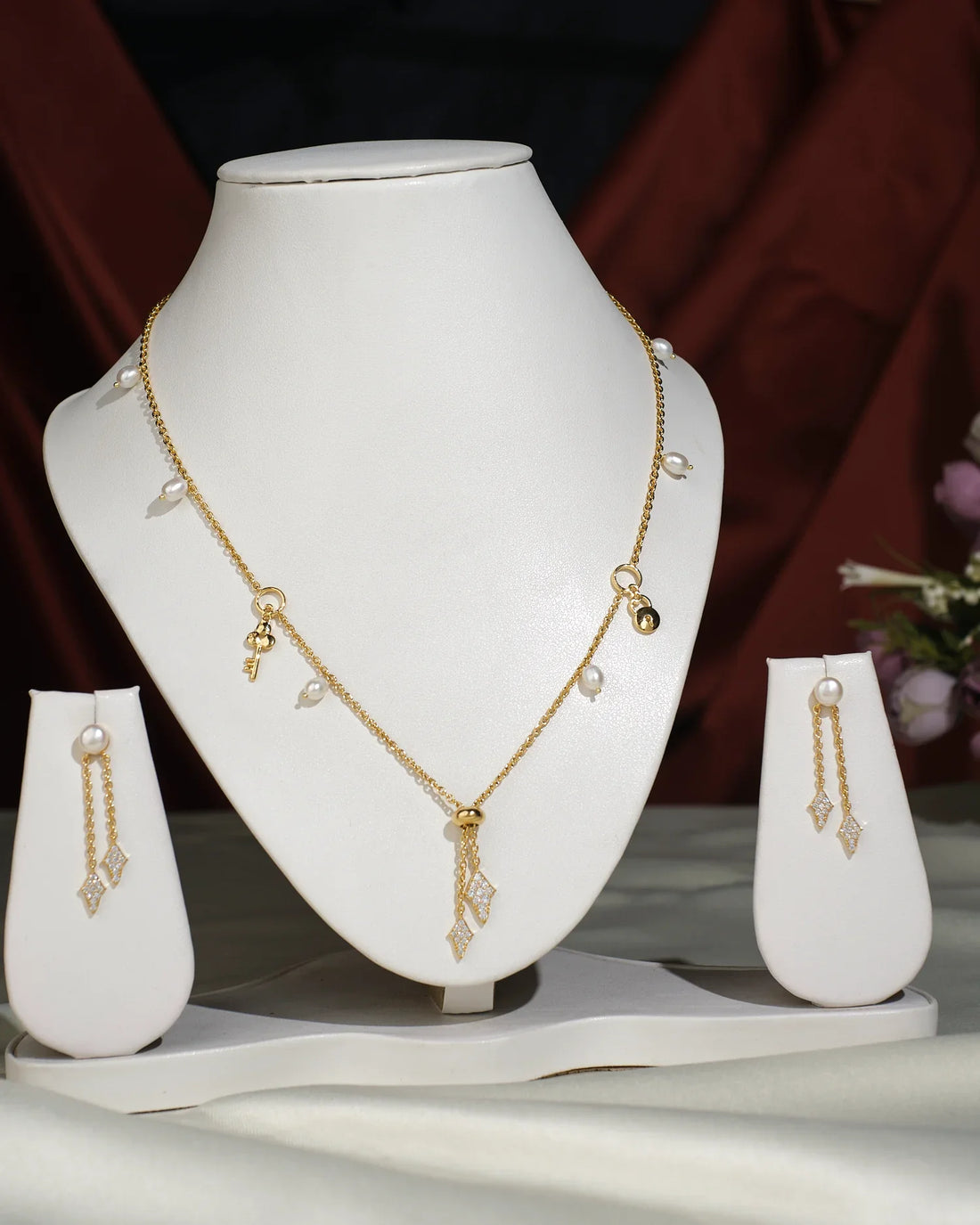 The Pretty Necklace Set by Chandrani Pearls India, featuring a gold necklace with key and lock charms and pearls, is showcased on a white stand alongside matching gold, diamond-shaped earrings on separate stands.