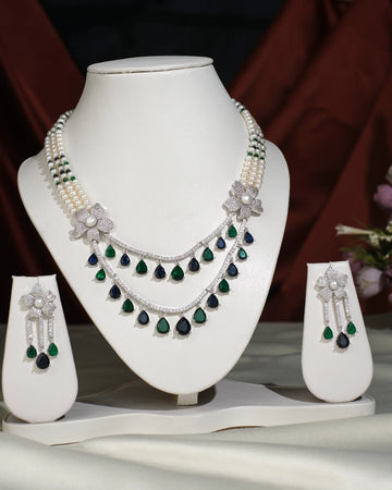 The Beautiful Ravishing Pearl Necklace Set by Chandrani Pearls India, featuring pearls, diamonds, and emeralds with matching earrings, is showcased on a white jewelry stand against a neutral background.