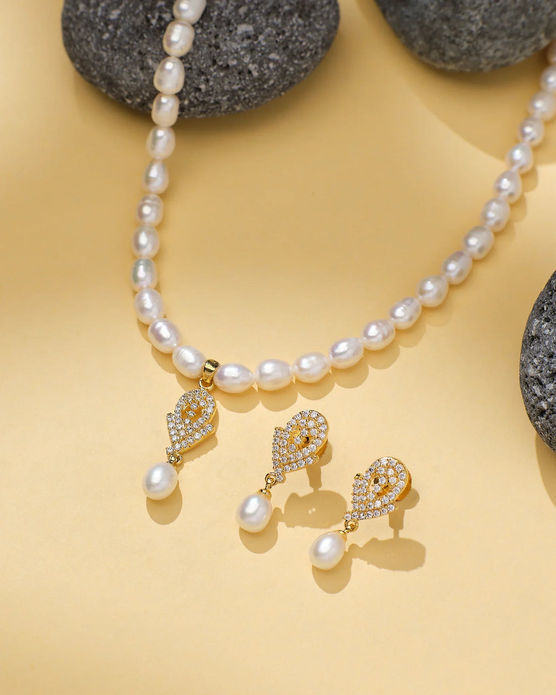 The Classic Beauty Pearl Set by Chandrani Pearls, featuring a pearl necklace and matching drop earrings with gold, CZ accents, and crystal details, is displayed on a beige surface with grey stones—ideal for elegant occasions.