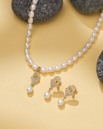 The Classic Beauty Pearl Set by Chandrani Pearls, featuring a pearl necklace and matching drop earrings with gold, CZ accents, and crystal details, is displayed on a beige surface with grey stones—ideal for elegant occasions.