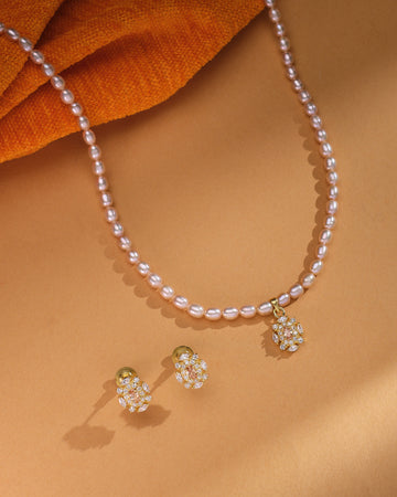 The Rosé Royale Necklace Set by Chandrani Pearls features a pearl necklace with a gold and diamond floral pendant and matching stud earrings, beautifully displayed on a tan surface with orange fabric—ideal for wedding jewelry.