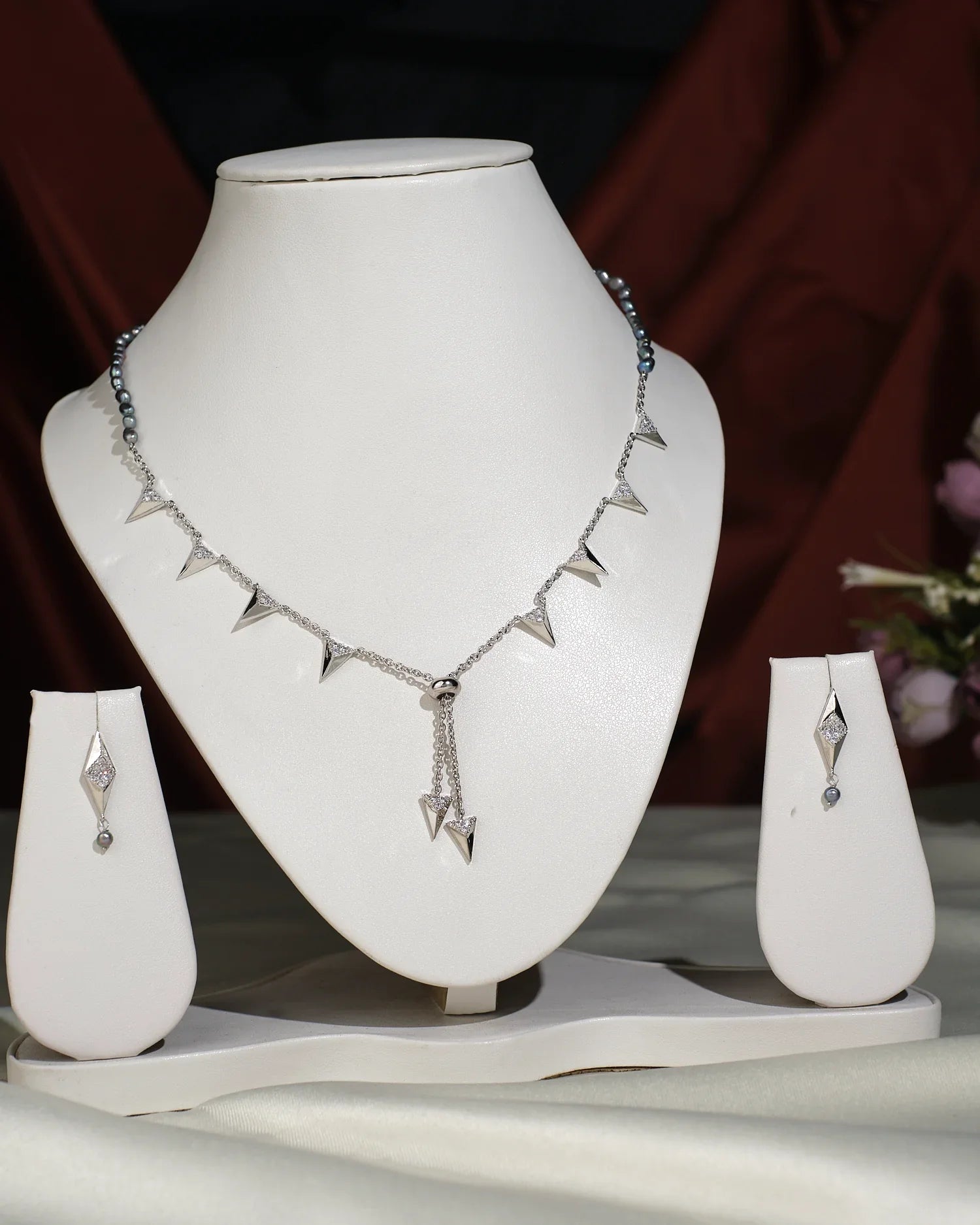 The Trendy Pearl Necklace Set by Chandrani Pearls India features a silver necklace with triangular pendants and matching earrings, showcased on white stands against contrasting fabric backgrounds, ideal for both casual and formal styles.