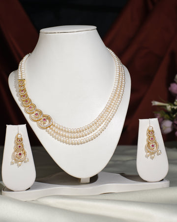 The Ravishing Pearl Necklace Set by Chandrani Pearls India, with a gold and pink accent, is displayed on a white stand alongside matching earrings on smaller stands—ideal for both formal and casual occasions.