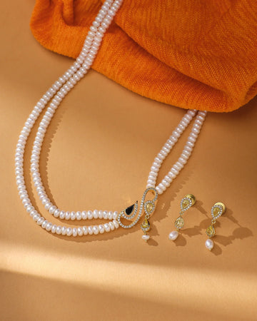 The Elegant & Classic Beauty Real Pearl Side Pendant Necklace & Earring Set by Chandrani Pearls, featuring real pearls and gold accents, is displayed on a beige surface with an orange fabric background.