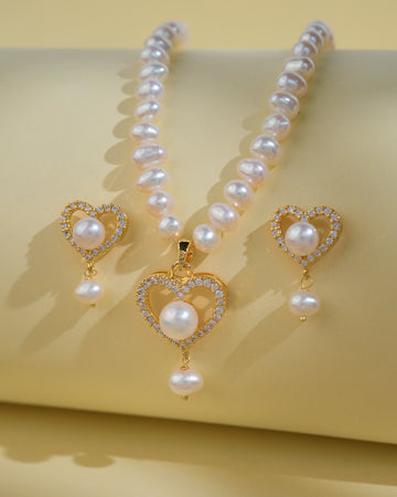 The Heart Pearl Necklace Set by Chandrani Pearls India features a pearl necklace with a gold heart-shaped pendant and matching earrings adorned with pearls, heart designs, and rhinestones, displayed on a light yellow surface.