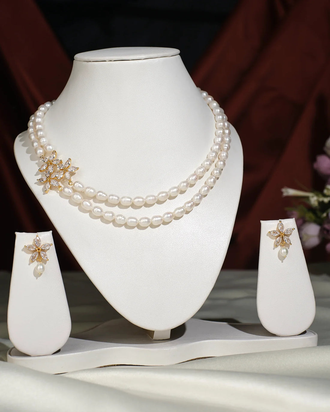 The Floral Pearl Necklace Set by Chandrani Pearls India features a double-stranded pearl necklace with a gold floral brooch and matching pearl and gold earrings, all displayed elegantly on a white stand.