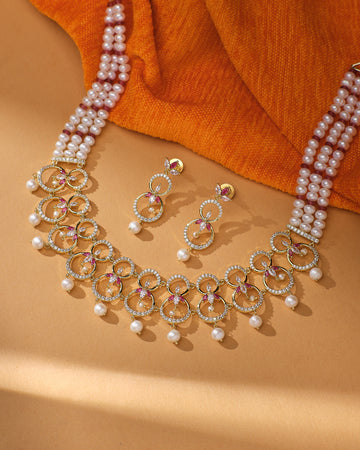 The Ravishing Real Pearl Bridal Jewelry Set by Chandrani Pearls features circular designs, pink accents, and pearl drops—ideal bridal jewelry. Displayed on a tan surface with an orange fabric backdrop.