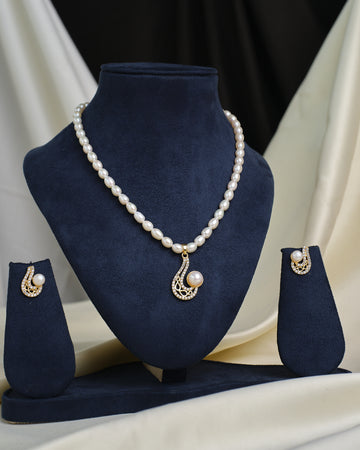 The Vintage-Inspired CZ Glam Set by Chandrani Pearls features a gold and pearl pendant necklace with matching earrings, displayed on a navy blue velvet stand against a cream and black background.