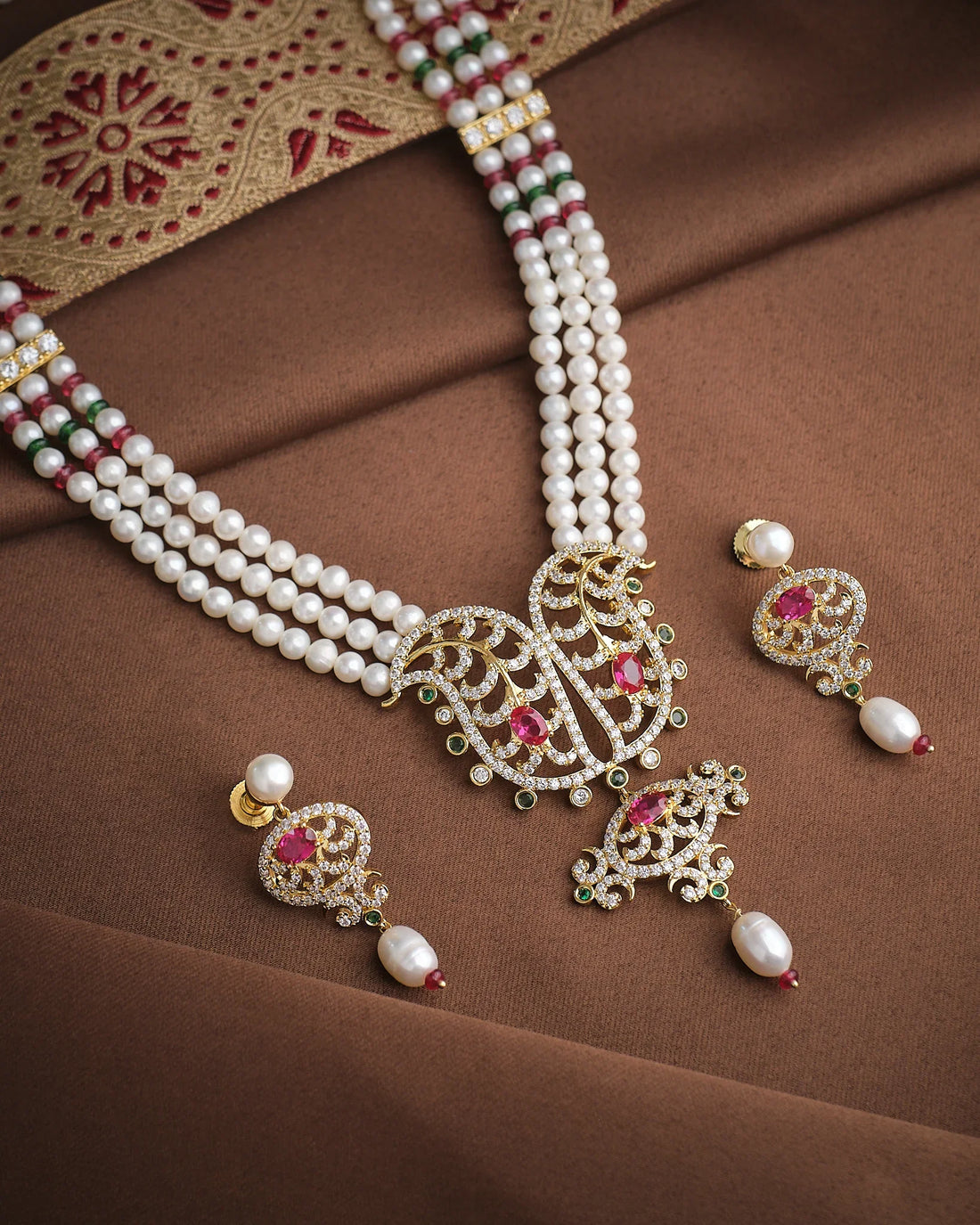 The Majestic Pearl & CZ Necklace Set by Chandrani Pearls features intricate designs, red and green gemstones, and matching earrings, sparkling brilliantly on brown embroidered fabric—perfect for any occasion.