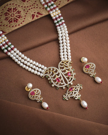 The Majestic Pearl & CZ Necklace Set by Chandrani Pearls features intricate designs, red and green gemstones, and matching earrings, sparkling brilliantly on brown embroidered fabric—perfect for any occasion.