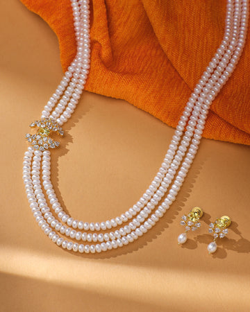 The Luxe Pearl Jewelry Set Glam Vibes Only by Chandrani Pearls features radiant pearls, a jeweled gold clasp, and matching floral gold earrings with pearl drops, elegantly displayed on a beige surface with an orange cloth.
