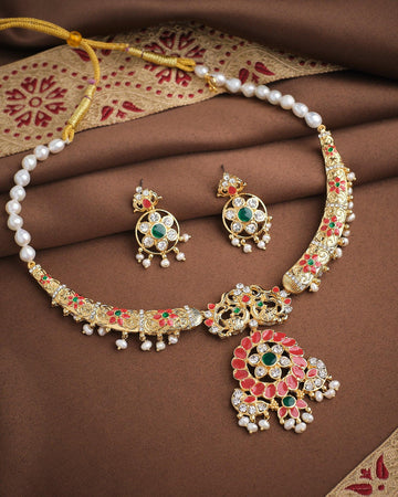 The Imperial Enamel Halo Pearl CZ Set by Chandrani Pearls features a gold and pearl necklace with matching earrings, adorned with green, red, and white enamel accents and haloed pearls, displayed on brown and gold patterned fabric.