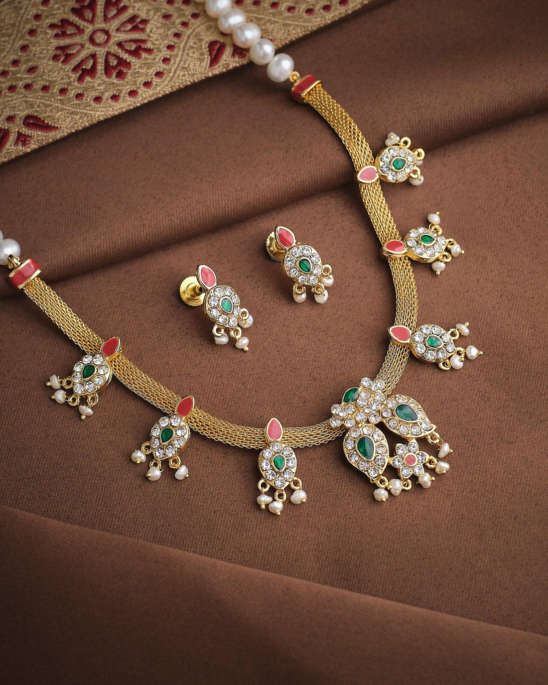 The Queen’s Radiant Pearl CZ Set by Chandrani Pearls features a gold necklace and matching earrings with green and white CZ stones, red accents, and pearl details, displayed on brown fabric with a patterned textile.