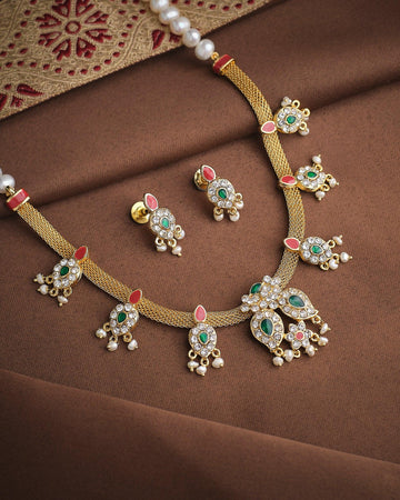 The Queen’s Radiant Pearl CZ Set by Chandrani Pearls features a gold necklace and matching earrings with green and white CZ stones, red accents, and pearl details, displayed on brown fabric with a patterned textile.