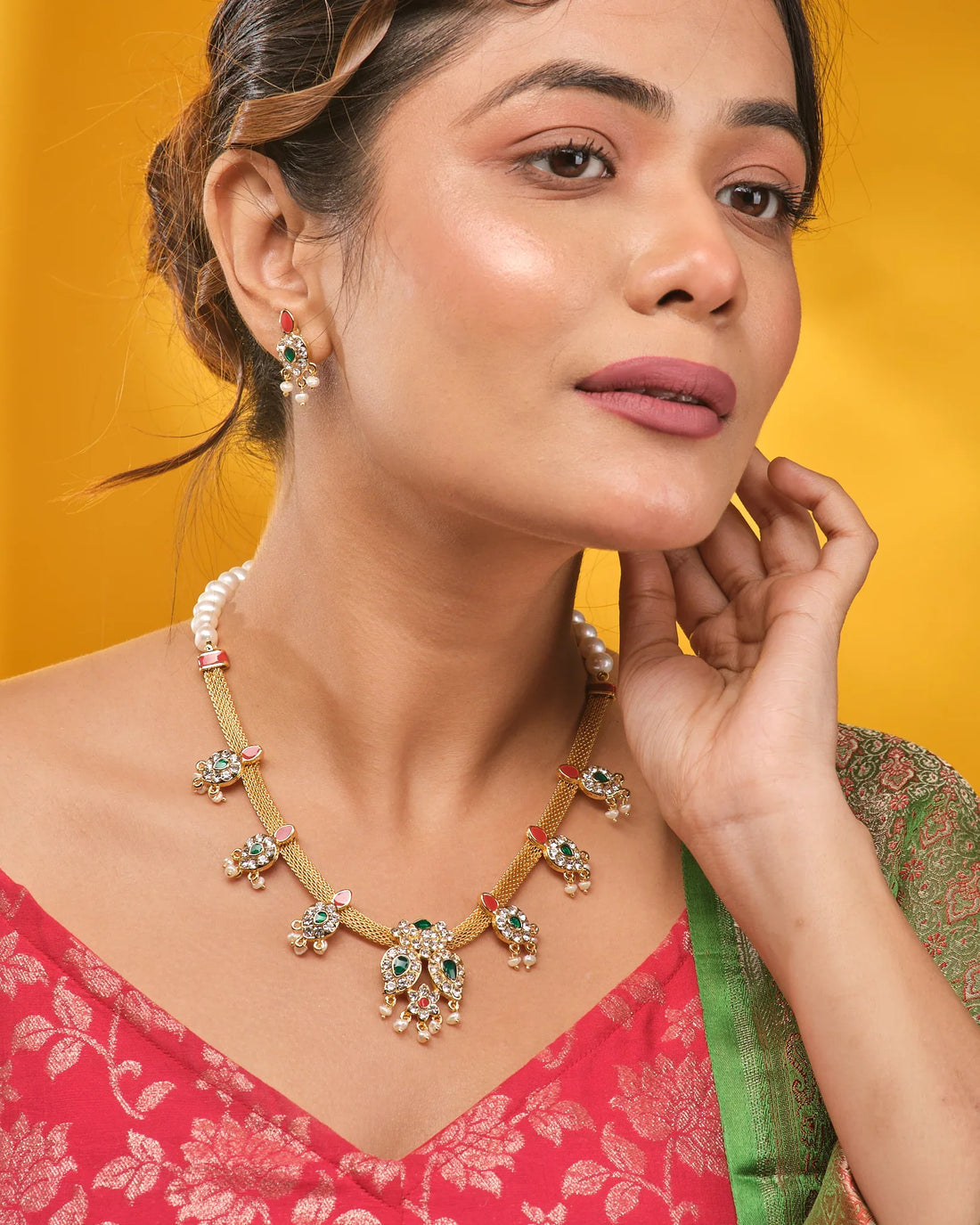 The Queen’s Radiant Pearl CZ Set by Chandrani Pearls features a gold necklace and matching earrings with green and white CZ stones, red accents, and pearl details, displayed on brown fabric with a patterned textile.